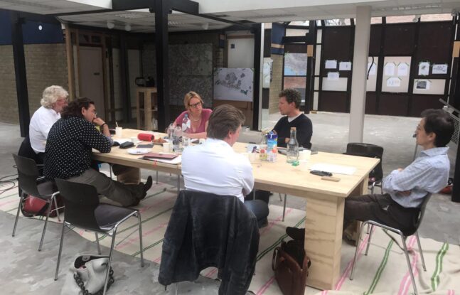 Zes mensen zitten aan rondom een tafel en overleggen met elkaar