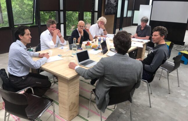 Zeven mensen zitten rondom een tafel met papieren en laptops voor zich te overleggen met elkaar