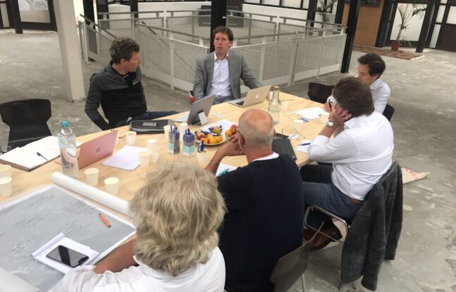 Zes mensen zitten aan een tafel met laptops voor zich en overleggen met elkaar