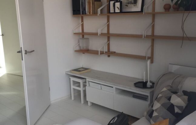 Een kleine woonkamer met aan de wand houtkleurige boekenplanken en op de voorgrond een witte bank
