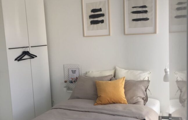 Een overwegend witte slaapkamer met tweepersoons bed in het midden en een kast aan de linkerkant