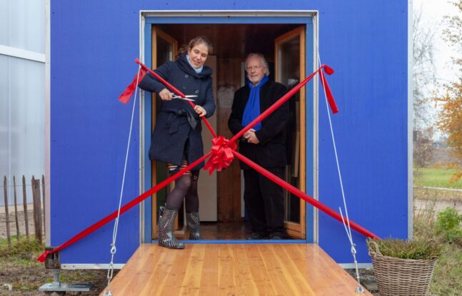 Tween mensen staan in een deuropening van een blauw tiny house met rood lint. Een persoon knipt het lint door.