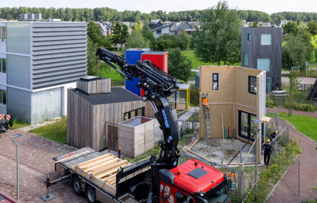Een kraanwagen staat met bouwmateriaal klaar bij een in opbouw zijnd tiny house